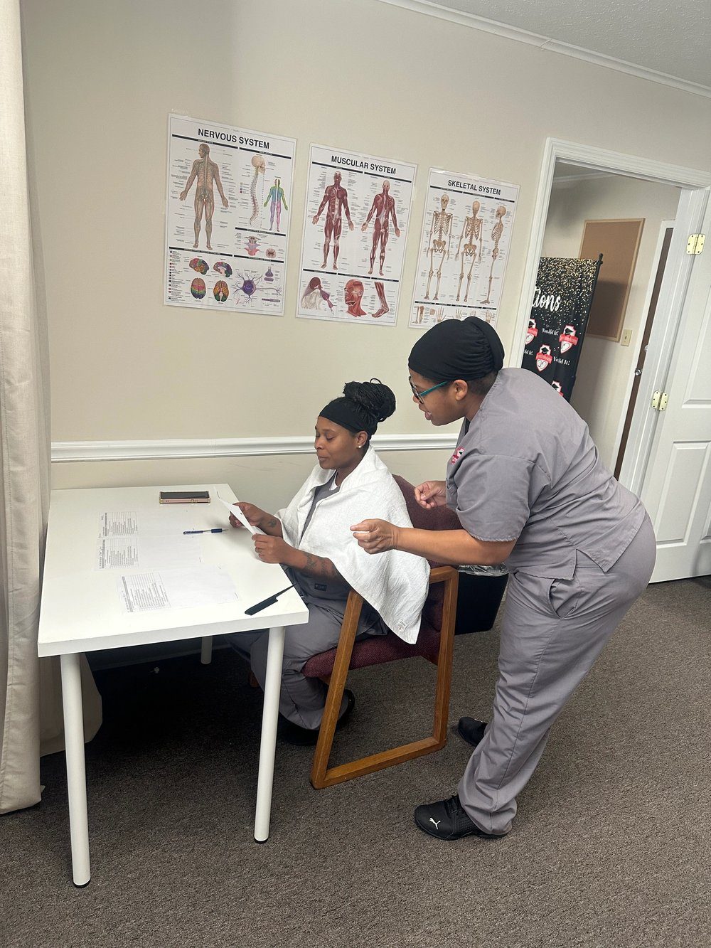 Two students in scrubs taking notes at a lab table beneath anatomy charts